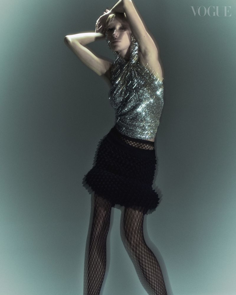 Blonde woman poses in a silver metallic top and black skirt with fishnet tights in a Vogue fashion editorial against a teal backdrop.