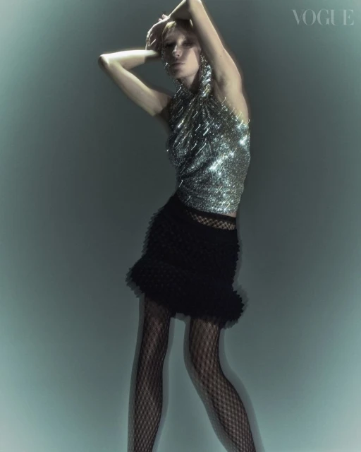 Blonde woman poses in a silver metallic top and black skirt with fishnet tights in a Vogue fashion editorial against a teal backdrop.