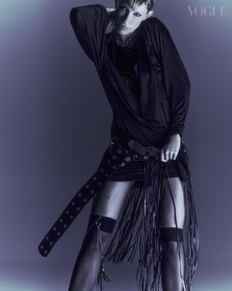 Fashion editorial shot of a woman in a long-sleeve top, mini dress with fringe, and thigh-high socks posing against a purple background.