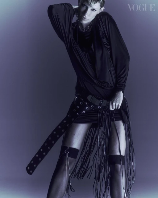 Fashion editorial shot of a woman in a long-sleeve top, mini dress with fringe, and thigh-high socks posing against a purple background.