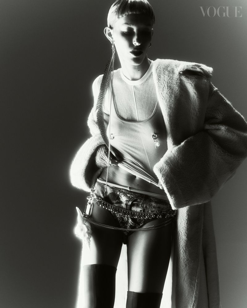Black and white fashion shot of a slim adult female wearing a coat, top, underwear, and boots, posing in a photo studio with a solid backdrop.