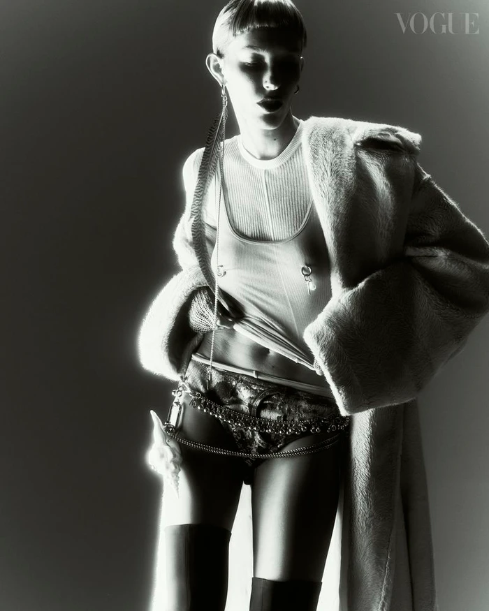 Model with short fringe wearing a textured coat over a ribbed top and dark patterned briefs against a plain light background