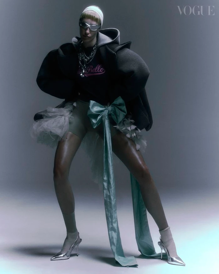 Model in futuristic sunglasses and hooded jacket with tulle tutu and silver heels against grey studio background
