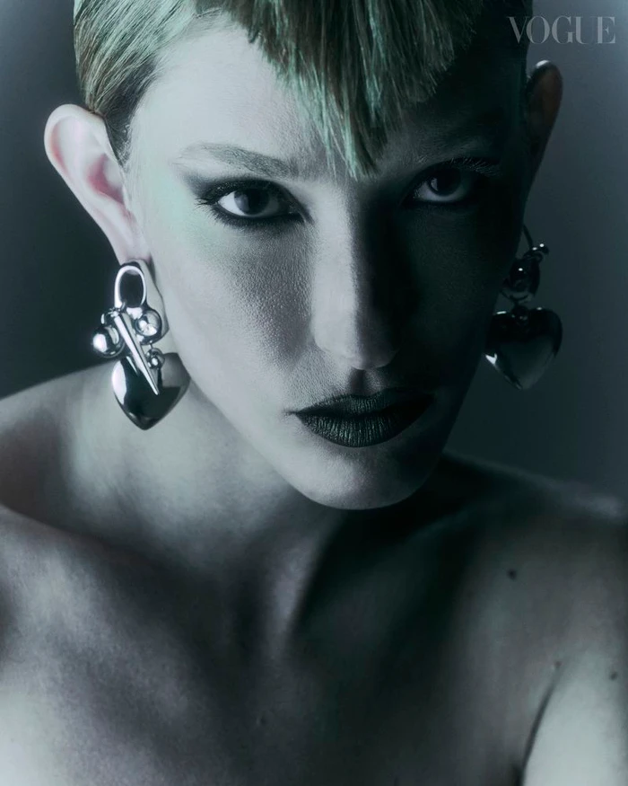 Close-up portrait of a model with short blonde hair and dark eye makeup wearing large silver heart earrings for Vogue Portugal.