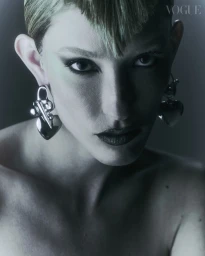 Close-up portrait of a model with short blonde hair and dark eye makeup wearing large silver heart earrings for Vogue Portugal.
