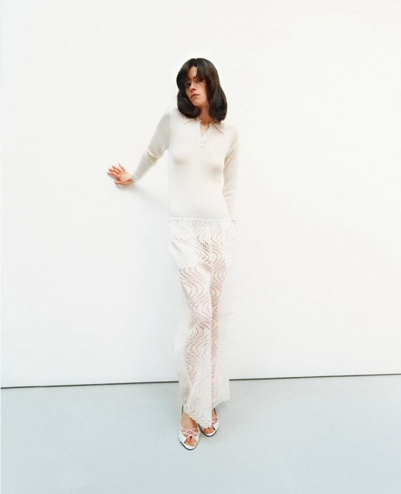 A brunette woman poses in a long-sleeve shirt and semi-sheer pants in a minimalist studio setting, looking elegantly at the camera.