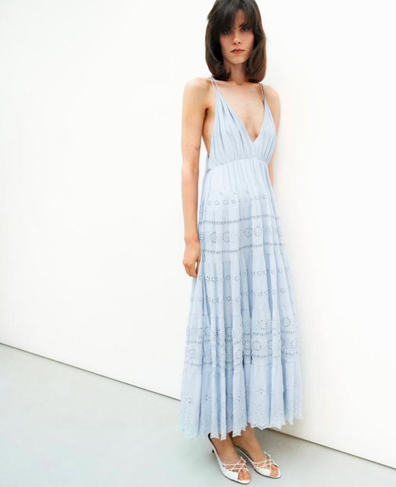 A woman poses indoors, wearing a light blue eyelet dress and white kitten heels against a white wall and floor backdrop.