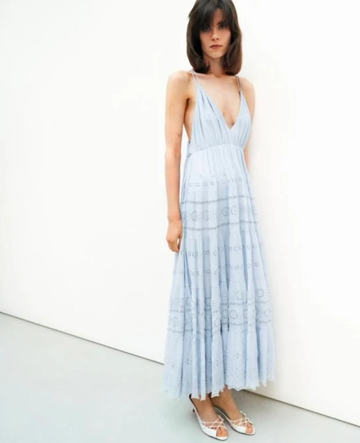 A woman poses indoors, wearing a light blue eyelet dress and white kitten heels against a white wall and floor backdrop.