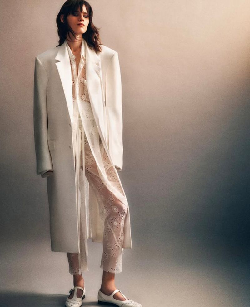 An adult female model poses in a white overcoat, eyelet trousers, and Mary Jane shoes, set against a graduated gray studio backdrop.