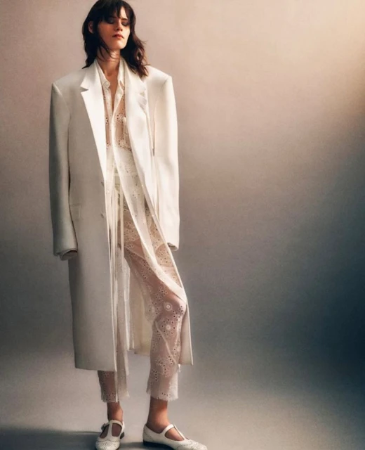 An adult female model poses in a white overcoat, eyelet trousers, and Mary Jane shoes, set against a graduated gray studio backdrop.