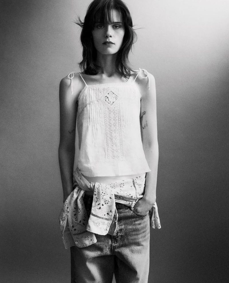 Monochrome portrait of a young adult woman with bangs wearing a white top and blue jeans, styled with a tied bandana around her waist.