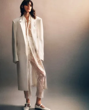 Model in a long white coat wearing sheer patterned trousers against a neutral studio backdrop