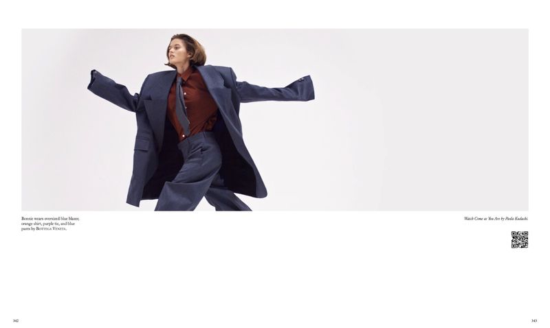 Fashion editorial shot of a female model in motion wearing an oversized blue suit, orange button-down shirt, and grey tie against a white background.