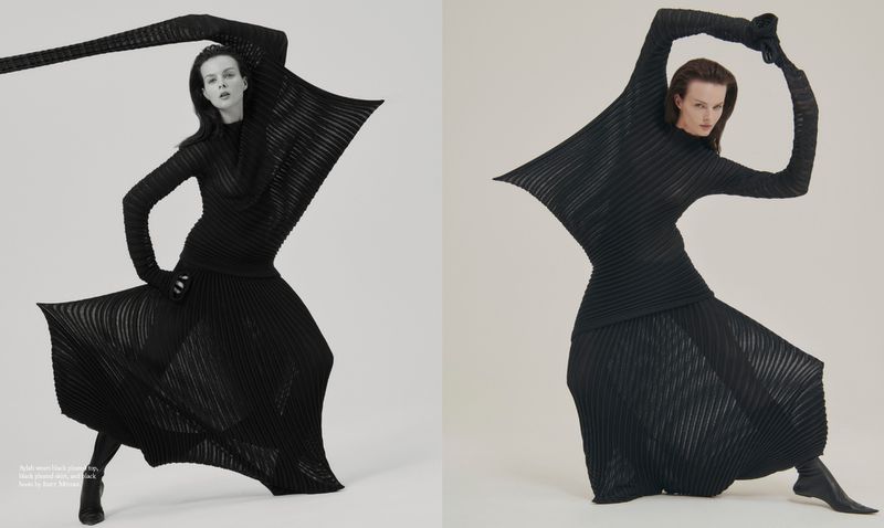 Fashion editorial shot of a model in a black pleated long-sleeve top, skirt, gloves, and boots, striking a dynamic pose on a white background.