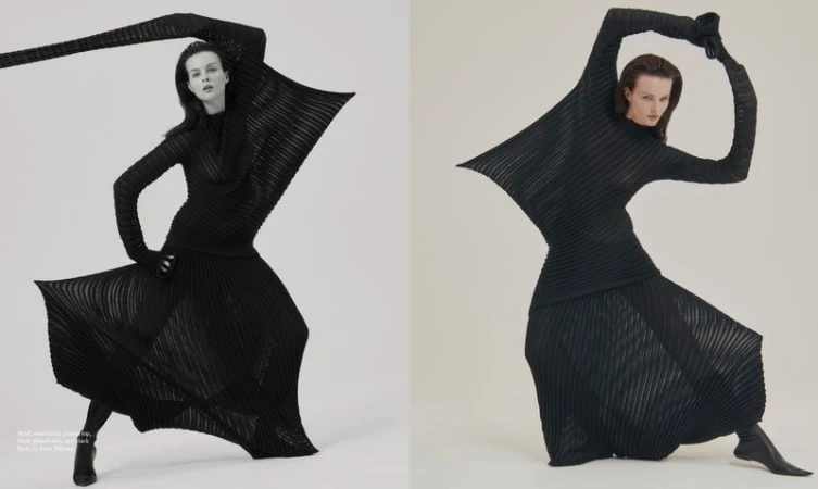 Fashion editorial shot of a model in a black pleated long-sleeve top, skirt, gloves, and boots, striking a dynamic pose on a white background.