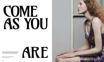 Profile view of a model in a purple sequin dress seated against a grey wall next to bold typography.