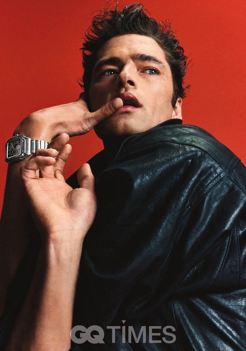 Portrait of a handsome adult male with dark hair wearing a black leather jacket and metal watch against a solid red backdrop.