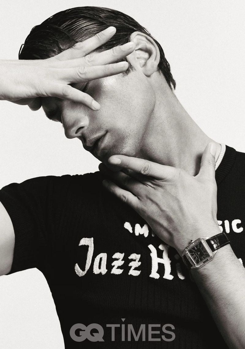 Monochrome portrait of a man in a black t-shirt holding his hand near his face, also wearing a watch with white background.