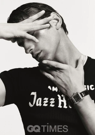 Monochrome portrait of a man in a black t-shirt holding his hand near his face, also wearing a watch with white background.