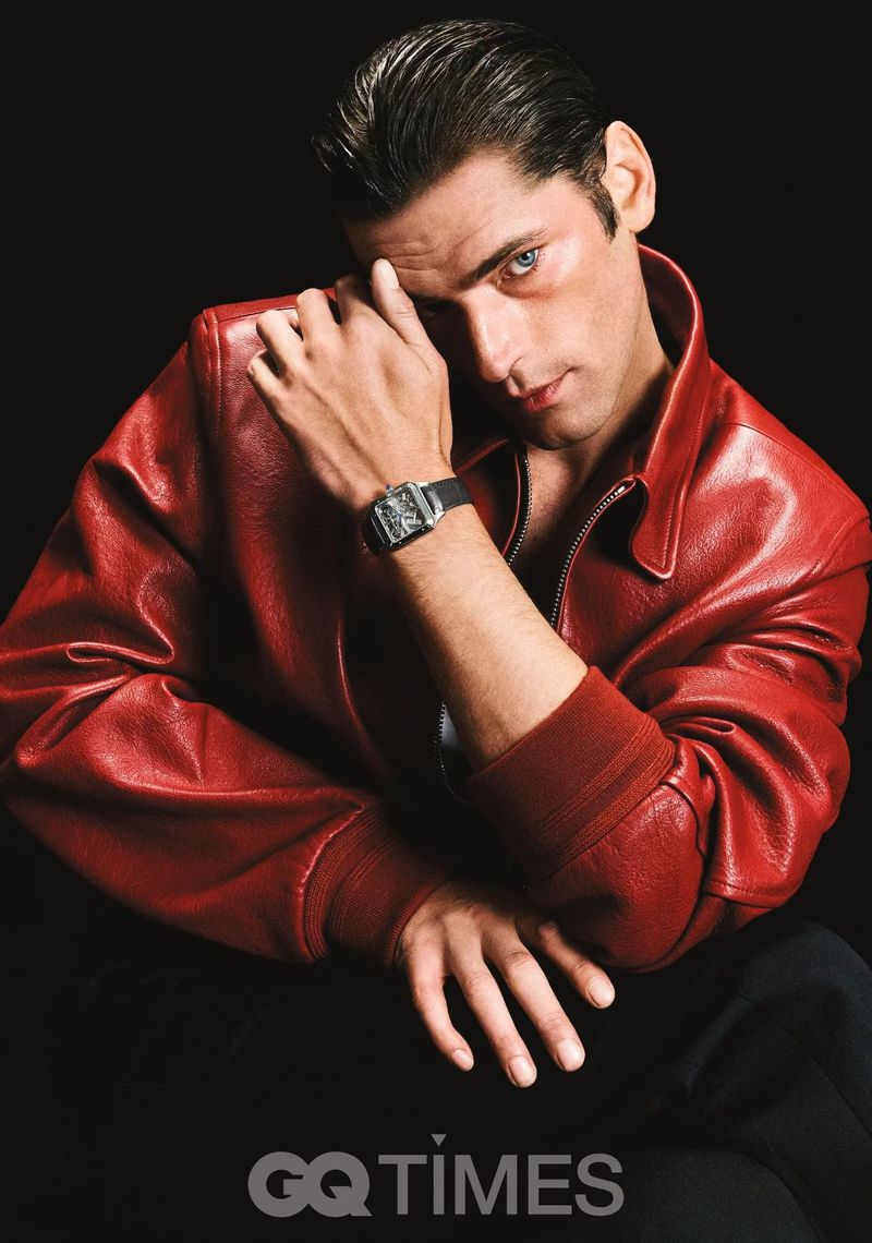 Portrait of a dark-haired man in a red jacket and watch, looking intently at the camera against a dark background in a studio setting.