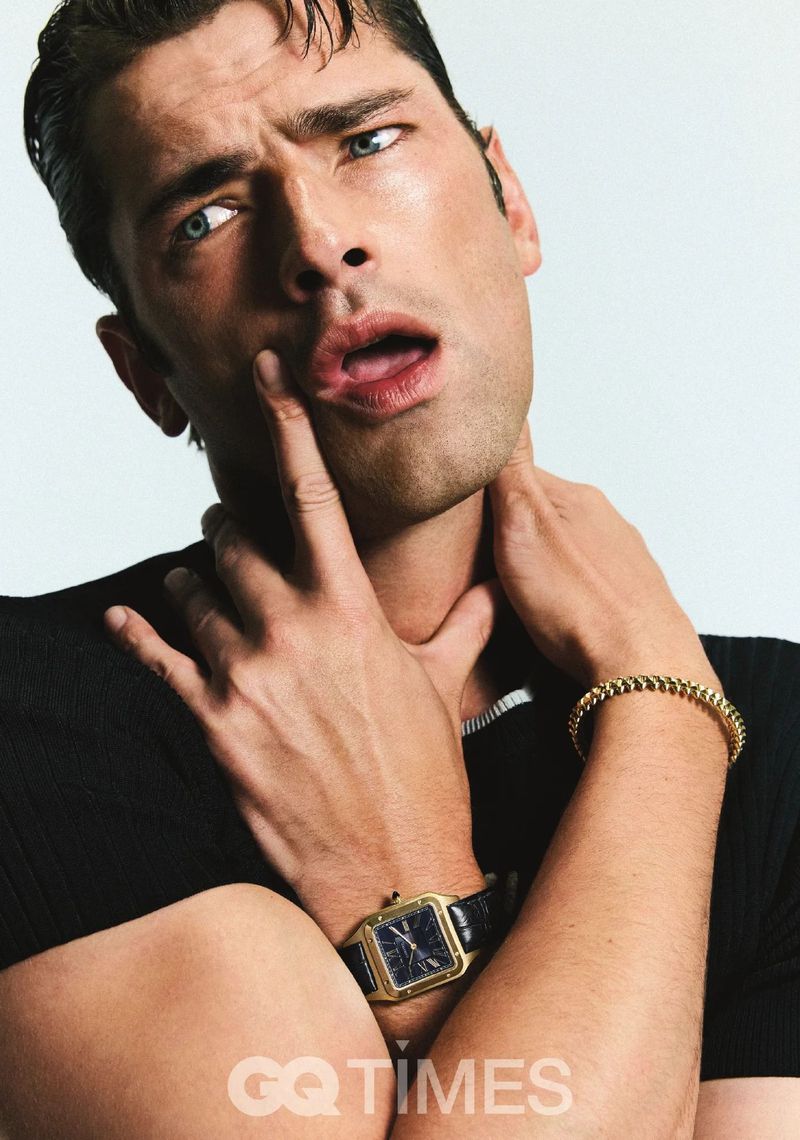 Portrait of a handsome man with light blue eyes wearing a black short-sleeve shirt, gold bracelet, and a watch against a white background.