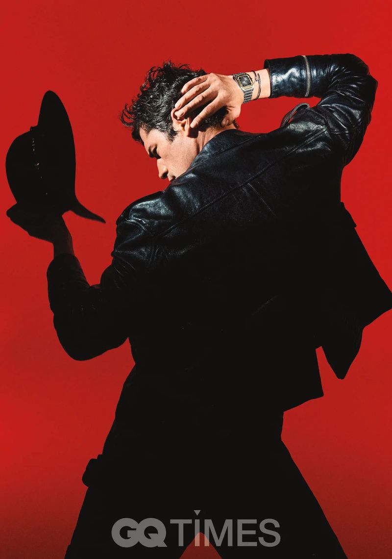 Adult male in black leather jacket holds hat against a red background, looking back over his shoulder in a fashion or editorial shot.