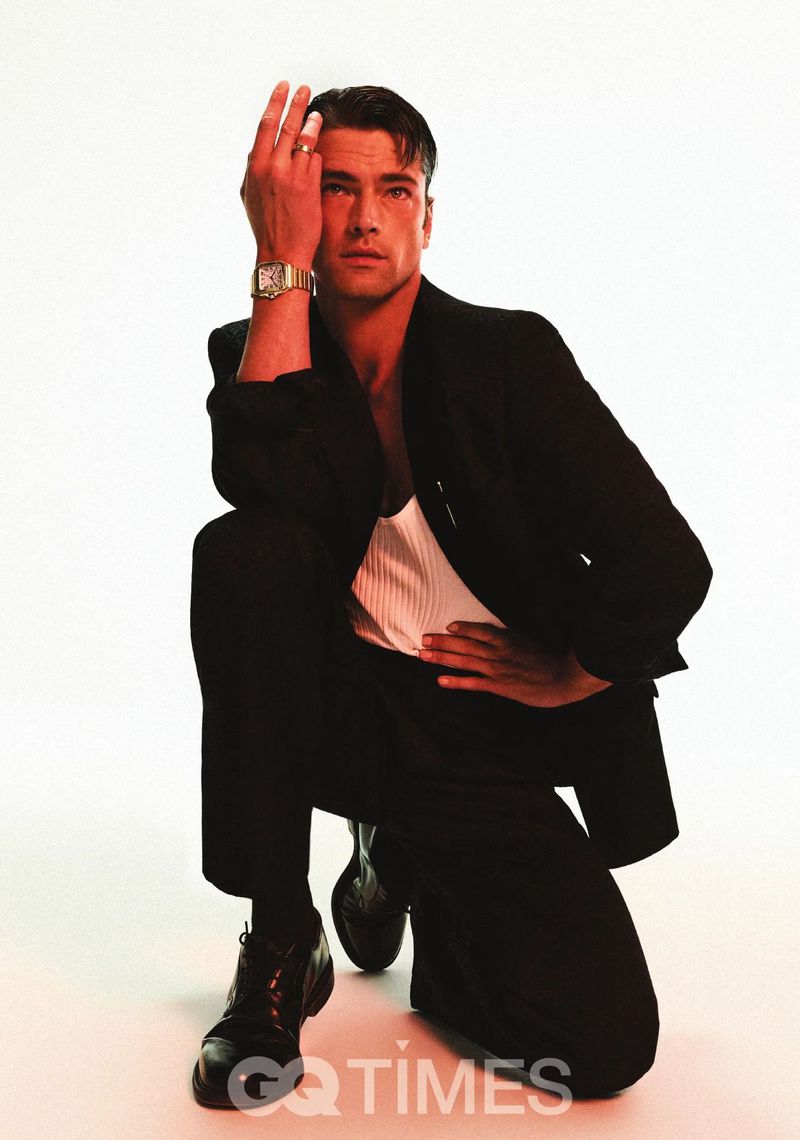 Portrait of an adult male crouched on white background wearing a black suit jacket, black pants, black socks, and black dress shoes.