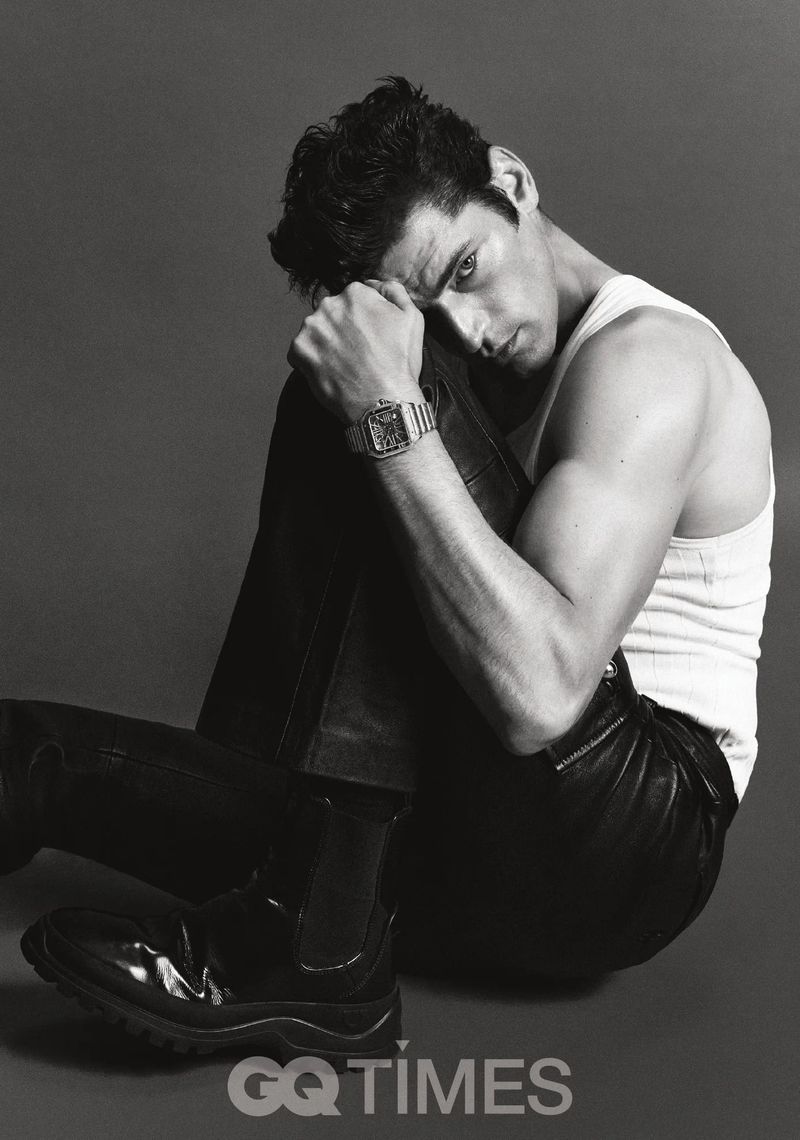 Monochrome portrait of a handsome adult man sitting in a photo studio wearing black pants, boots, and a white tank top.