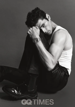 Monochrome portrait of a handsome adult man sitting in a photo studio wearing black pants, boots, and a white tank top.