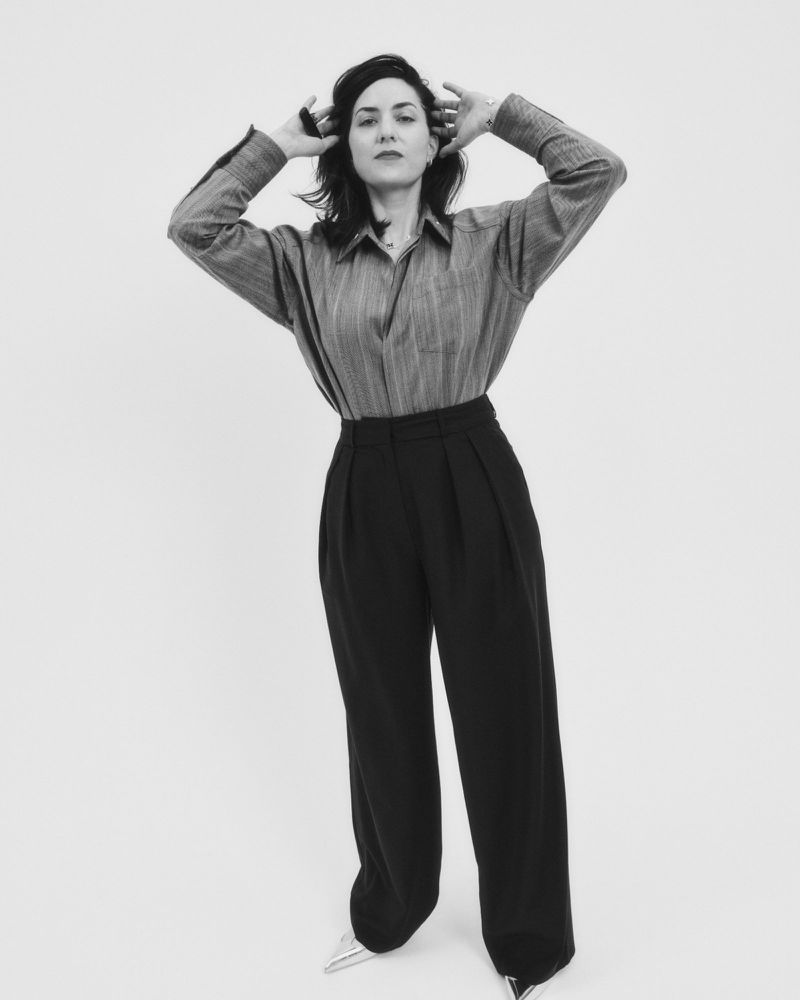A striking black and white portrait of a woman in an oversized button-down shirt and wide-leg pants, set against a clean white backdrop.