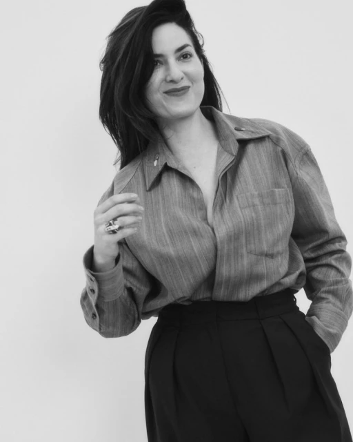 Monochrome portrait of a woman in casual attire, featuring a button-down shirt, dark pants, and a ring, set against a bright background.