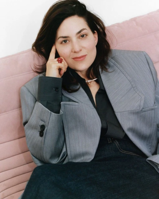 Portrait of adult woman with dark hair wearing a gray blazer and dark shirt, reclining on a pink sofa against a white wall.