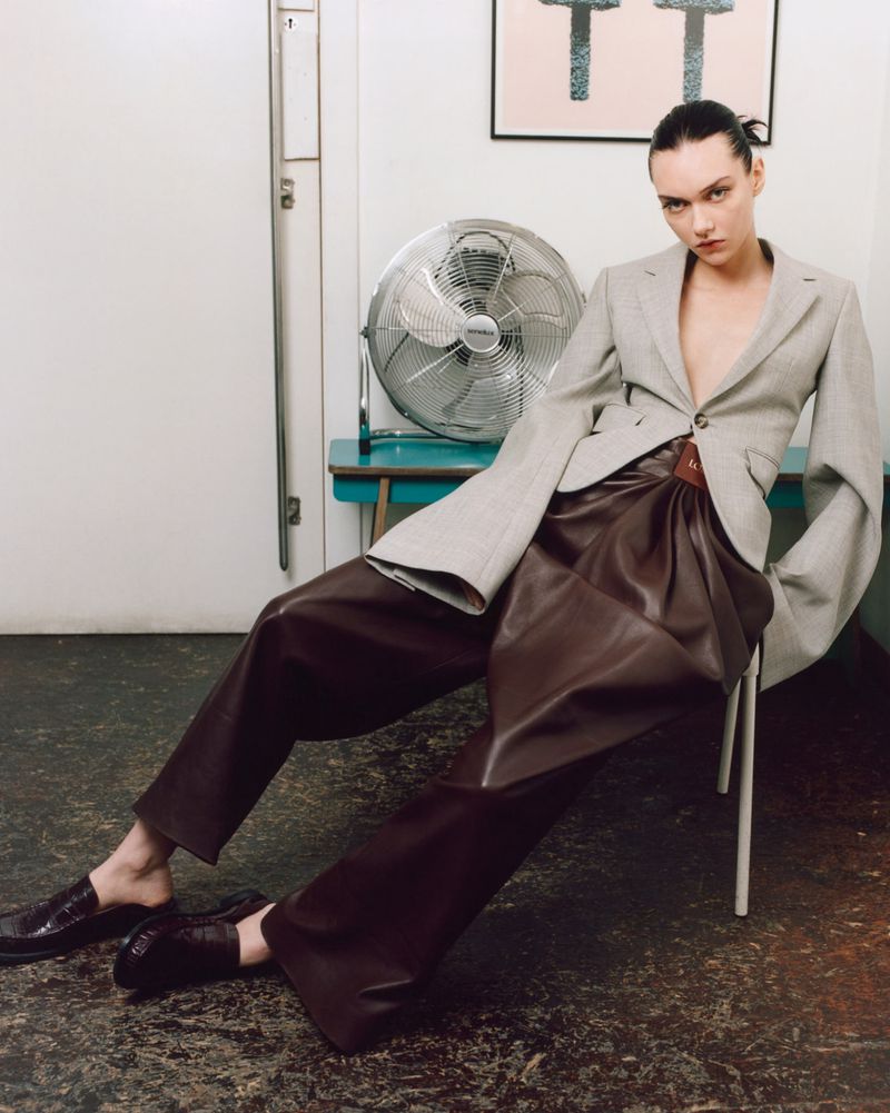Woman posing in oversized blazer and dark brown leather pants in a minimalist indoor setting with fan and abstract art.