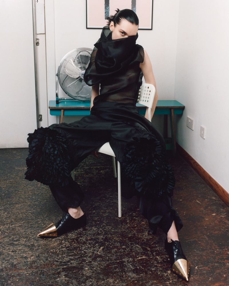 Editorial fashion shot of a person with a sheer black ruffled top and large black ruffled pants, seated on a white chair indoors.