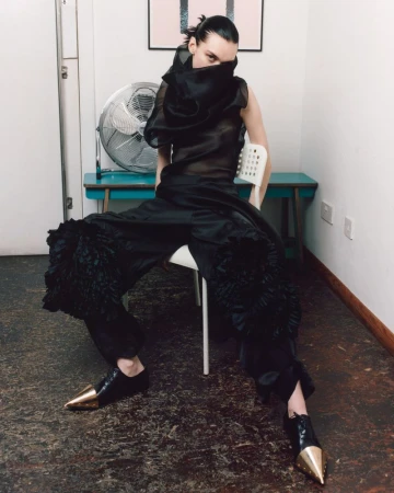 Editorial fashion shot of a person with a sheer black ruffled top and large black ruffled pants, seated on a white chair indoors.