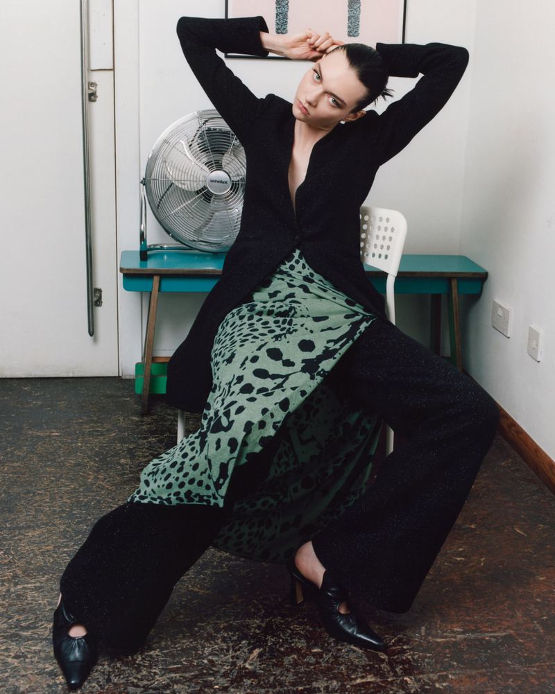 A model in a black jacket and patterned skirt poses on a chair in a white room with a vintage fan and blue table, creating a fashion editorial.