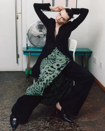 A model in a black jacket and patterned skirt poses on a chair in a white room with a vintage fan and blue table, creating a fashion editorial.