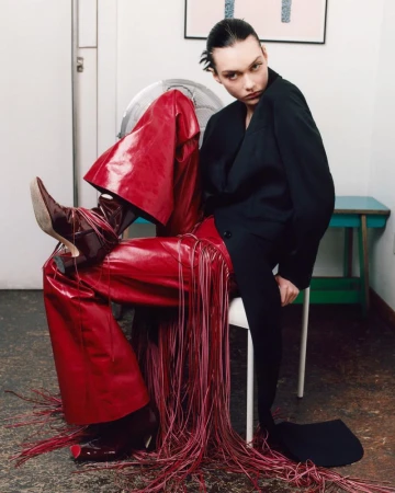 A fashion-forward individual in red leather pants with fringe sits on a white chair indoors against a white wall, showcasing unique style.