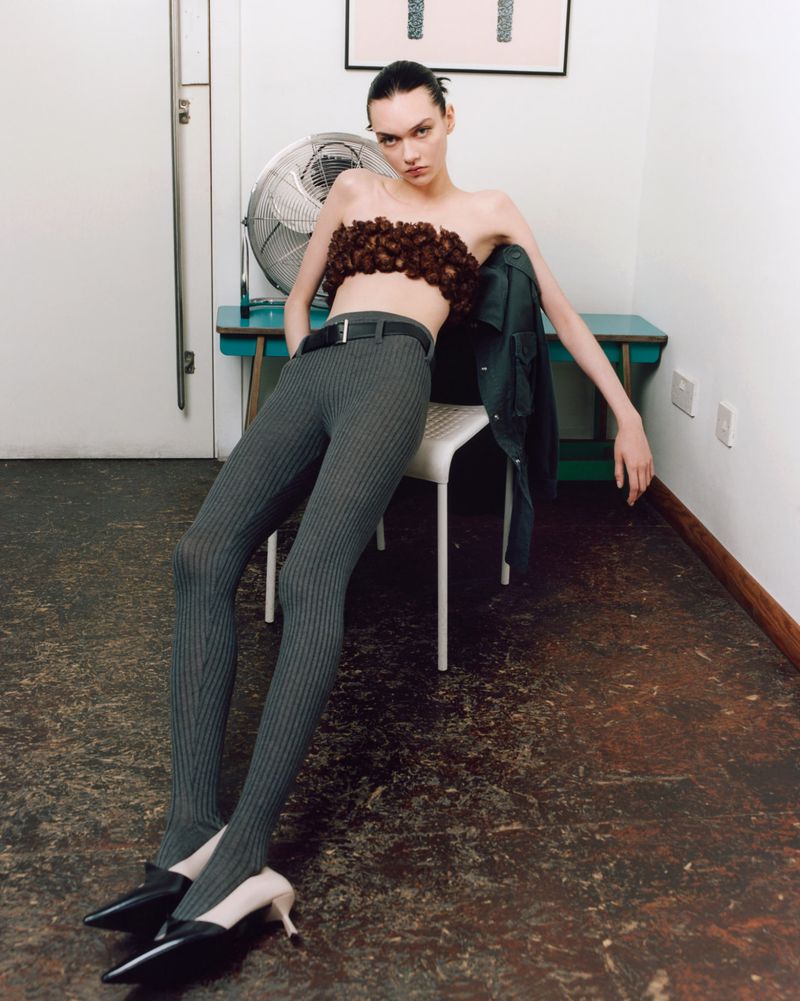 A slender woman with a brown-flower tube top, and grey pinstripe tights poses in a sparse indoor setting, next to a vintage desk fan.