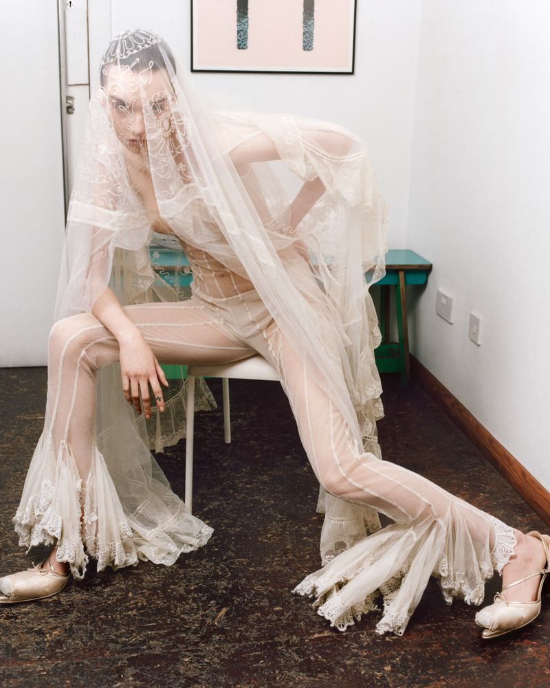 A stylish fashion shot features a model in a sheer, lace-trimmed outfit, seated indoors with an abstract art piece in the background.