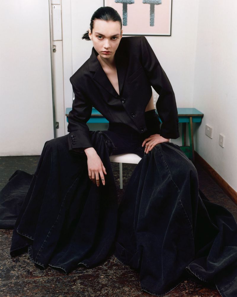 A striking fashion shot of a young woman in an oversized black blazer and flowing pants sitting indoors on a white chair.