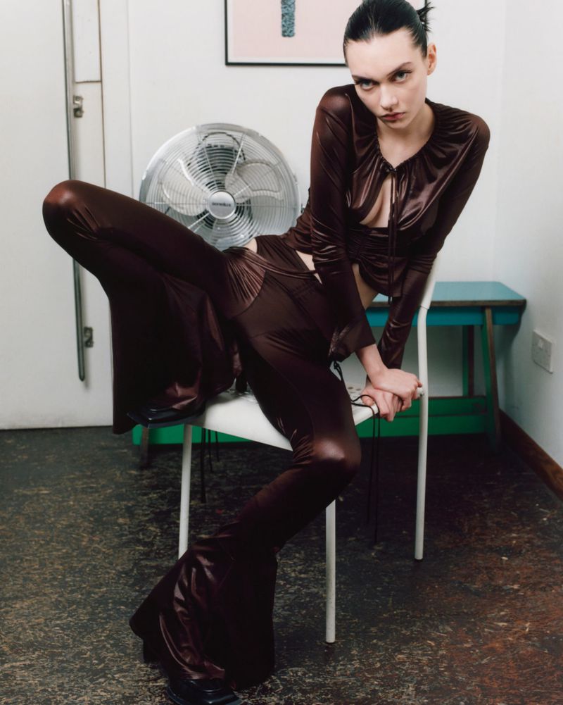 Brunette model in a brown satin outfit, sitting on a white chair with a fan in the background, posing for a fashion shot indoors.