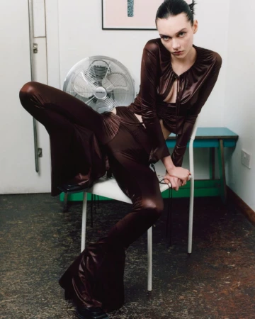 Brunette model in a brown satin outfit, sitting on a white chair with a fan in the background, posing for a fashion shot indoors.