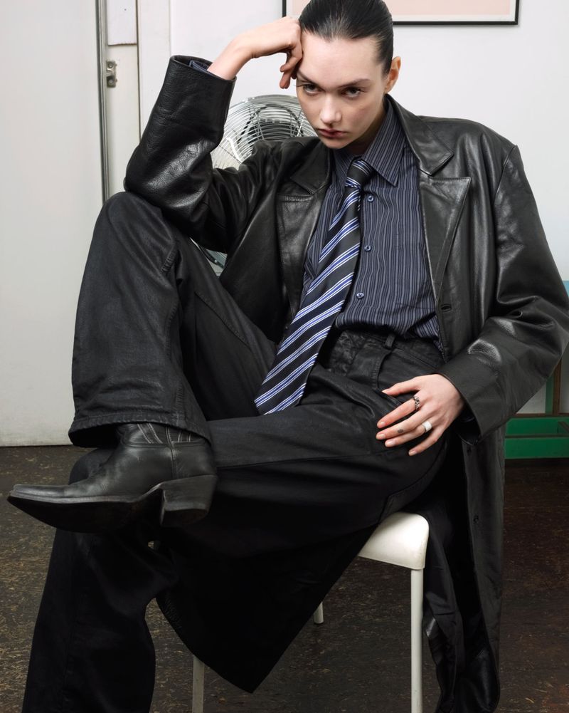 Brunette woman poses in a long black leather coat, black pants, and boots while sitting in a simple indoor setting.