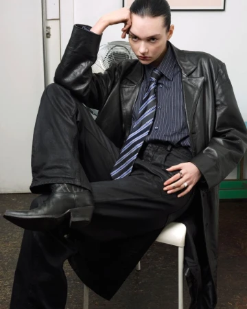 Brunette woman poses in a long black leather coat, black pants, and boots while sitting in a simple indoor setting.