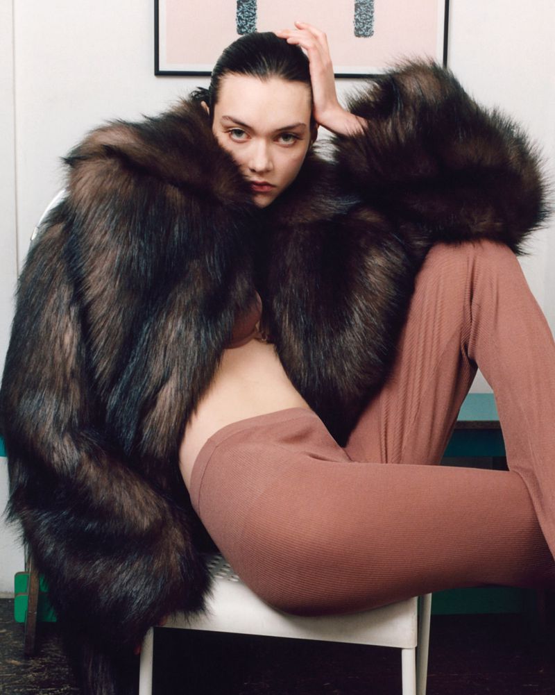 Woman in fur coat seated on a chair in a photo studio. Model poses while wearing brown pants and a brown fur coat.