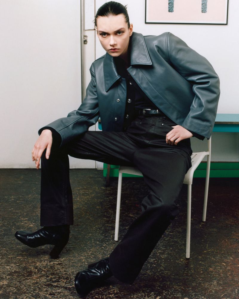 A fashionable woman with slicked-back hair poses indoors, wearing a leather jacket, dark pants, and heeled boots, seated on a white chair.
