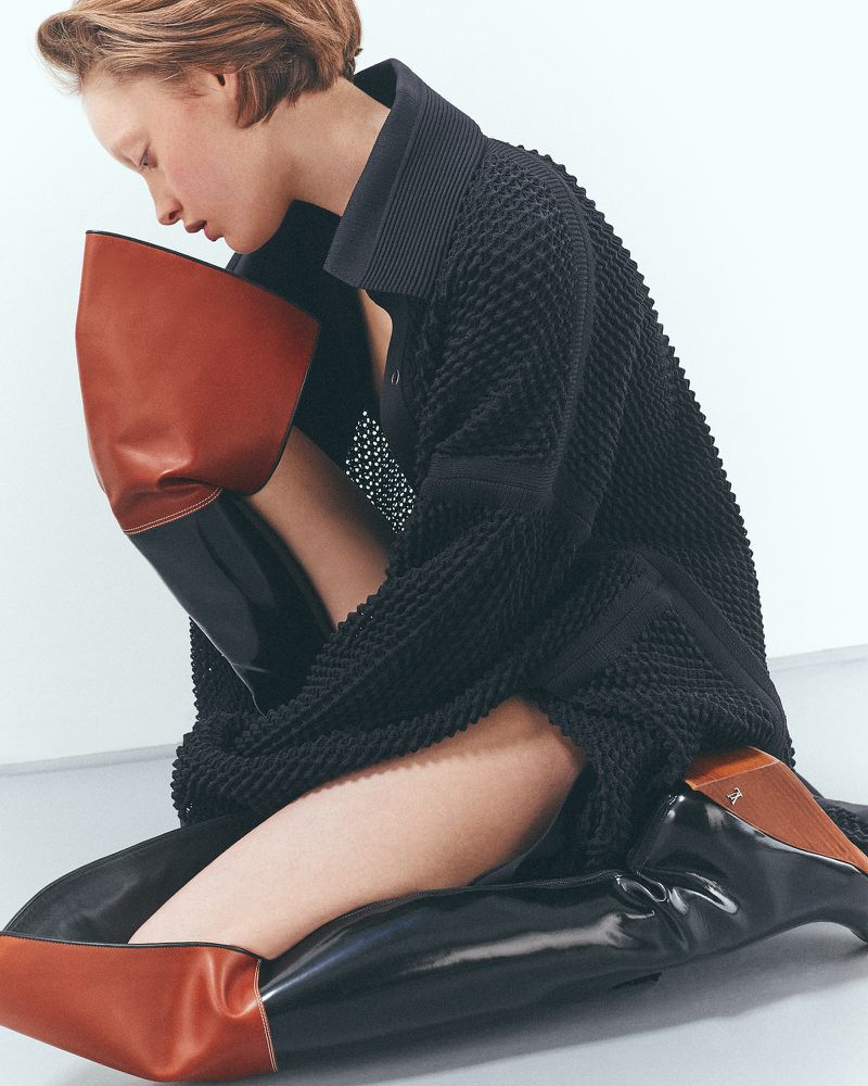 Fashion model in a textured black dress poses indoors with brown and black boots against a white background in a studio fashion editorial.