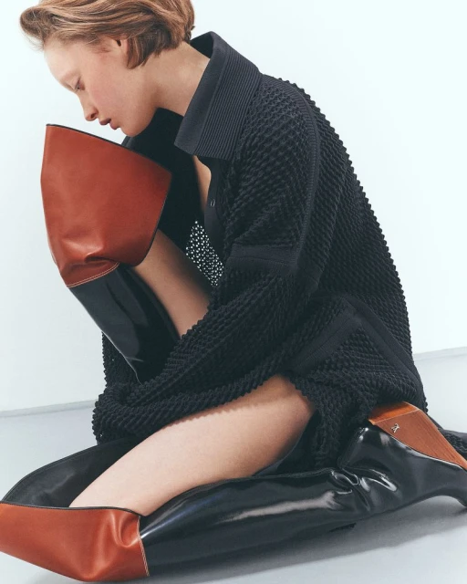 Fashion model in a textured black dress poses indoors with brown and black boots against a white background in a studio fashion editorial.