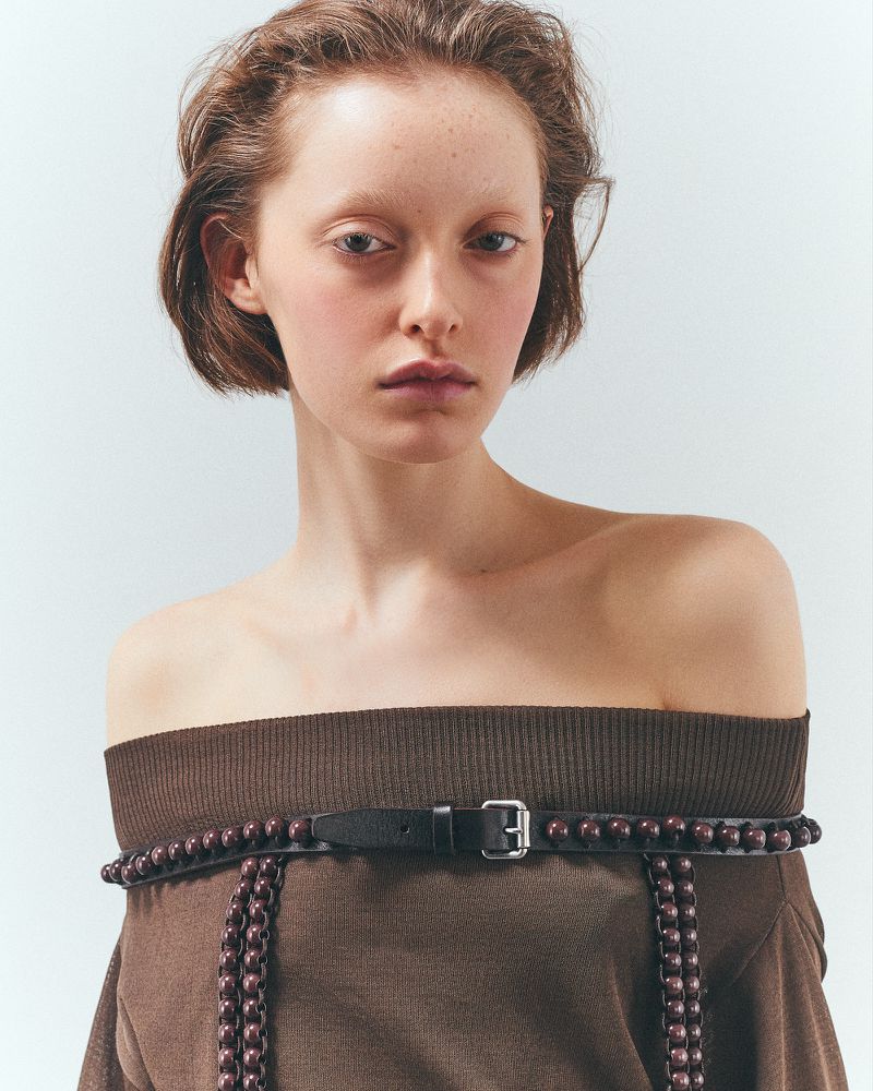 Portrait of an adult female fashion model wearing a brown knit off-the-shoulder top with a beaded belt against a plain background.